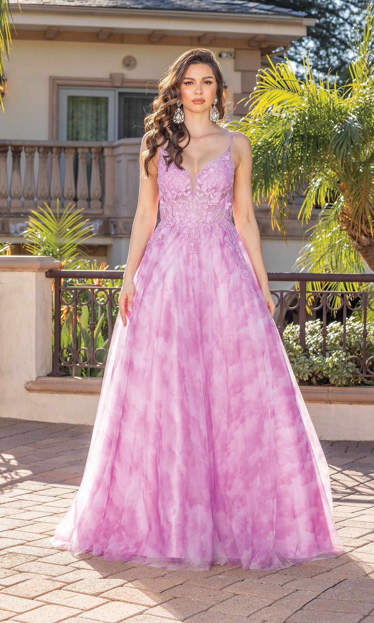 A woman stands outdoors in front of a railing wearing the Tie-Dye Prom Ball Gown with Sheer Sides. This affordable formal dress features a long, sleeveless design, perfect for special occasions. Plants and a building are in the background.